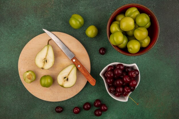 Vista superior de frutas como medio corte pera y ciruela con cuchillo en la tabla de cortar y tazones de cereza y ciruela sobre fondo verde