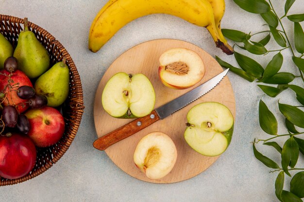 Vista superior de frutas como medio melocotón cortado y manzana con un cuchillo en la tabla de cortar y una canasta de pera uva melocotón con plátano y hojas sobre fondo blanco.