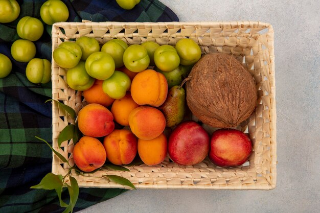 Vista superior de frutas como melocotón, albaricoque, ciruela, coco, pera en canasta sobre tela escocesa y fondo blanco.