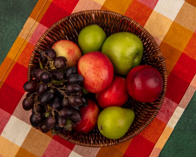 Foto gratuita vista superior de frutas como melocotón manzana pera uva en canasta sobre tela escocesa y sobre fondo verde