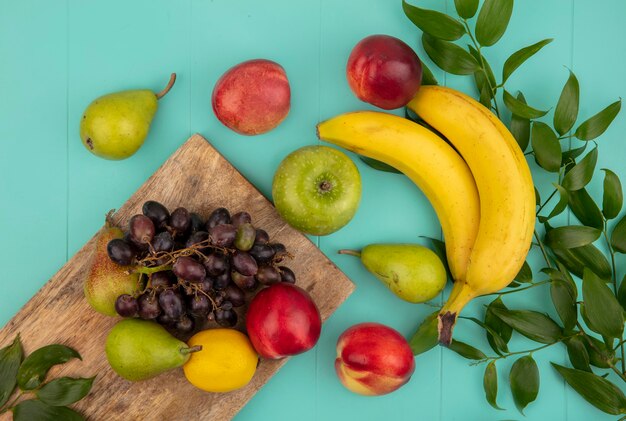 Vista superior de frutas como melocotón pera limón uva en tabla de cortar con plátano manzana y hojas sobre fondo azul.