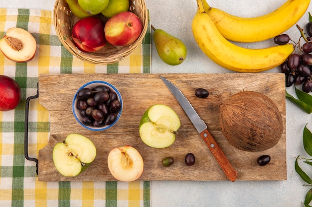 Foto gratuita vista superior de las frutas como la mitad de la manzana cortada, melocotón y uva de coco con un cuchillo en la tabla de cortar y una canasta de manzana melocotón en tela escocesa con uva plátano sobre fondo blanco.