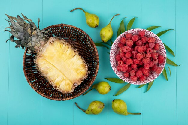 Vista superior de frutas como la mitad de piña en la canasta y el tazón de frambuesa con duraznos y hojas sobre superficie azul