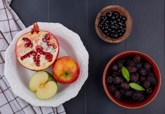 Vista superior de frutas como mitades de granada y manzana con manzana entera en plato sobre tela escocesa y cuencos de endrinas y moras en superficie negra