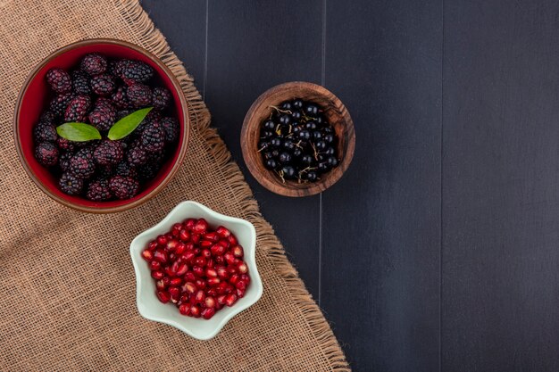 Vista superior de frutas como moras y bayas de granada en tazones de tela de saco con tazón de bayas endrinas en superficie negra