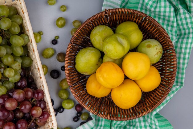 Vista superior de frutas como nectacots pluots verdes en canasta sobre tela escocesa y uvas en canasta y sobre fondo gris