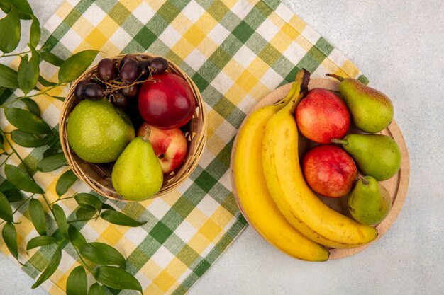 Vista superior de frutas como pera manzana melocotón uva plátano en canasta y en tabla de cortar con hojas sobre tela escocesa y fondo blanco.