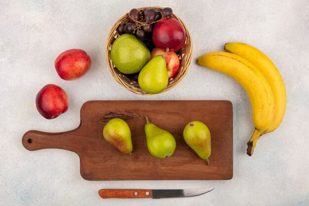 Vista superior de frutas como peras en tabla de cortar y canasta de melocotón uva manzana pera con plátanos y cuchillo sobre fondo blanco.