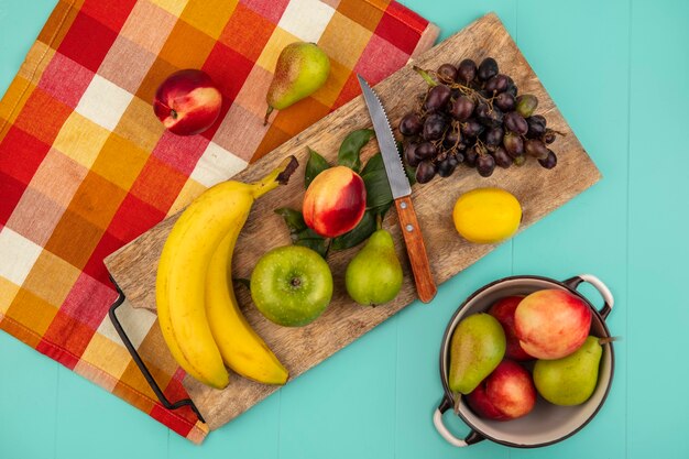 Foto gratuita vista superior de frutas como plátano manzana melocotón pera limón uva con cuchillo y hojas sobre tabla de cortar en tela escocesa con olla de melocotón pera sobre fondo azul