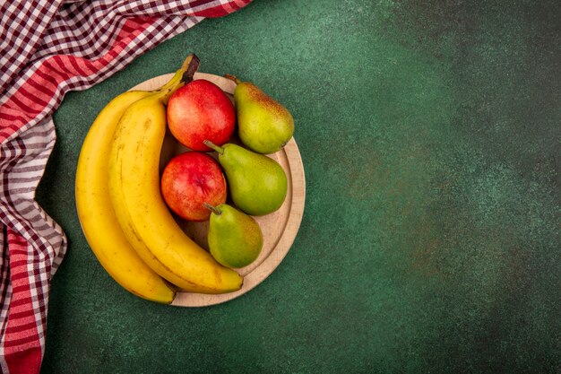 Vista superior de frutas como plátano melocotón pera en tabla de cortar con tela escocesa sobre fondo verde con espacio de copia