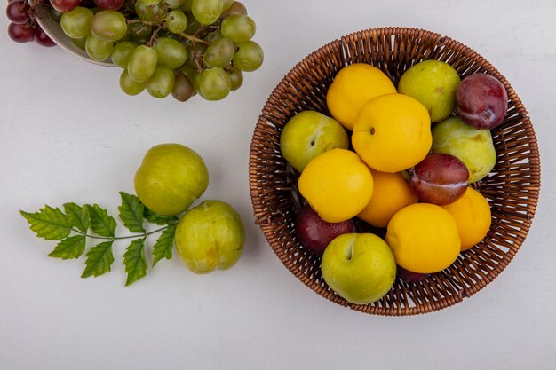 Vista superior de frutas como pluots nectacots en canasta con uva en tazón y pluots verde sobre fondo blanco.