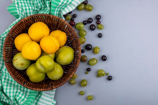 Vista superior de frutas como pluots verdes y nectacots en canasta sobre tela escocesa con bayas de uva sobre fondo gris con espacio de copia