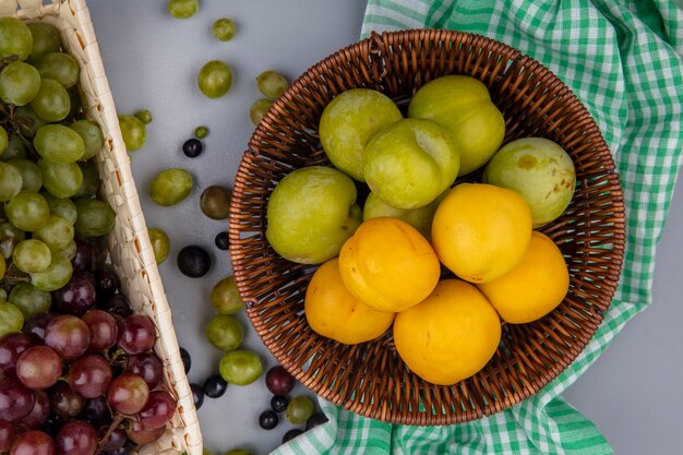 Vista superior de frutas como pluots verdes y nectacots en canasta sobre tela escocesa y canasta de uvas con bayas de uva sobre fondo gris