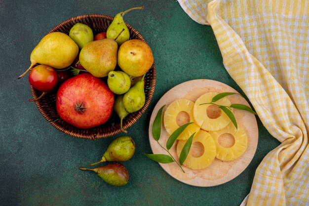 Vista superior de frutas como rodajas de piña en la tabla de cortar con granada, durazno y ciruela en la cesta sobre la superficie verde