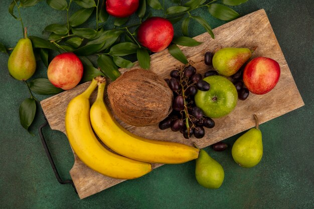 Vista superior de frutas como uva melocotón pera manzana plátano coco en tabla de cortar con hojas sobre fondo verde