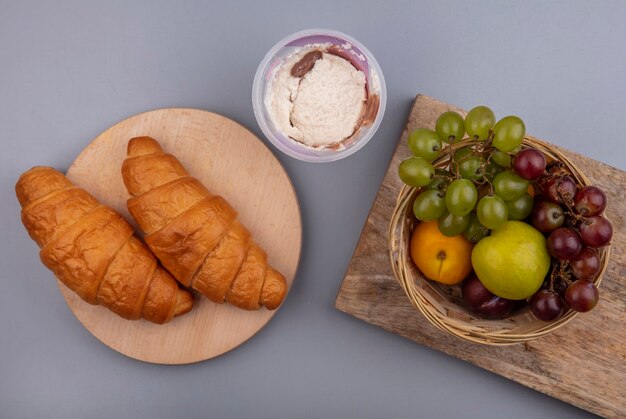 Vista superior de frutas como uva nectacot pluot en canasta con croissants en tablas de cortar sobre fondo gris