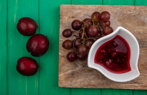 Vista superior de frutas como uva y pluots con mermelada de frambuesa en tabla de cortar sobre fondo verde