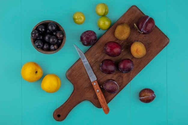 Foto gratuita vista superior de las frutas cortadas y enteras y un cuchillo en la tabla de cortar con ciruelas nectacots y un tazón de bayas de uva sobre fondo azul.