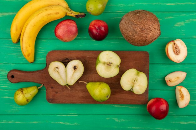 Vista superior de frutas enteras y medio cortadas como manzana pera en tabla de cortar con coco de plátano melocotón sobre fondo verde