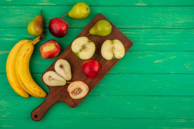 Vista superior de frutas enteras y medio cortadas como pera manzana melocotón en tabla de cortar con plátanos sobre fondo verde con espacio de copia