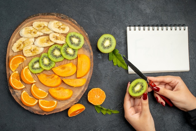 Vista superior de frutas frescas en una bandeja de madera para picar una rodaja de kiwi junto al cuaderno sobre fondo oscuro