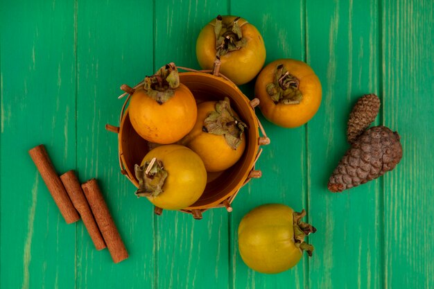 Vista superior de frutas frescas de caqui en un balde con ramas de canela en una mesa de madera verde