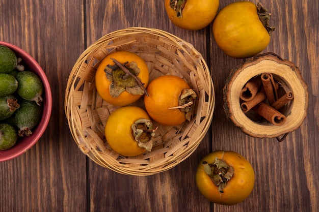 Vista superior de frutas frescas de caqui en un balde con ramas de canela en un tarro de madera sobre un fondo de madera