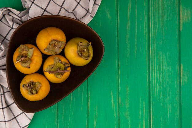 Vista superior de frutas frescas de caqui en un recipiente sobre un paño marcado sobre una mesa de madera verde con espacio de copia