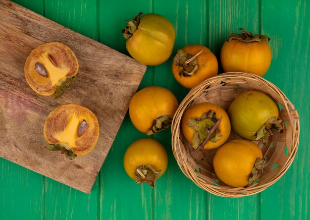 Vista superior de frutas frescas de caqui en una tabla de cocina de madera con frutas de caqui en un balde sobre una mesa de madera verde