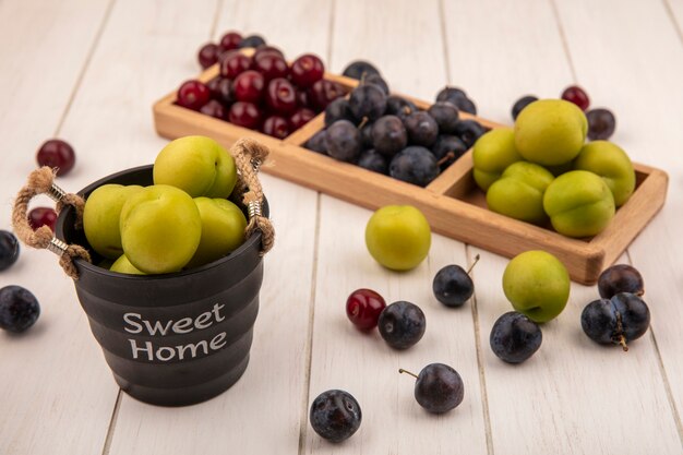 Vista superior de frutas frescas como cereza verde, cerezas rojas y endrinas en una bandeja de madera dividida con ciruela cereza verde en una canasta sobre un fondo blanco.