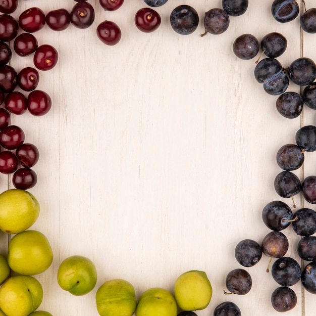 Vista superior de frutas frescas como ciruelas cereza verde y endrinas aisladas sobre un fondo de madera blanca con espacio de copia