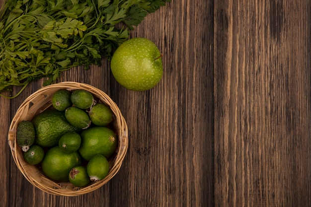 Vista superior de frutas frescas como feijoas y limas en un balde con manzanas y perejil aislado en una superficie de madera con espacio de copia