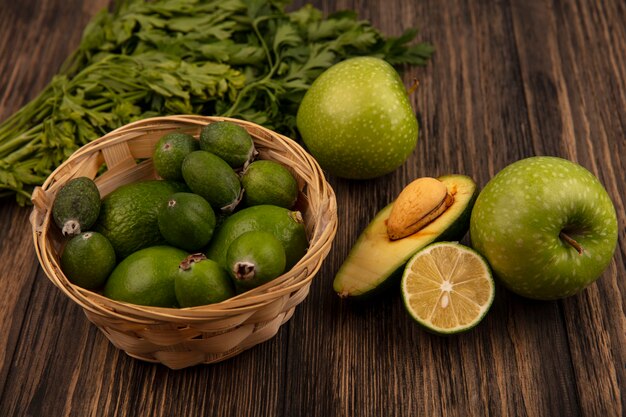 Vista superior de frutas frescas como feijoas y limas en un balde con medio aguacate y limón con manzanas y perejil aislado en una superficie de madera