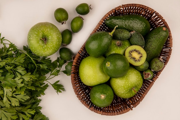 Vista superior de frutas frescas como kiwi, manzanas, aguacates, limas y feijoas en un balde sobre una pared blanca.