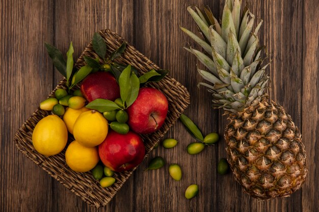 Vista superior de frutas frescas como manzanas, limones y kinkans en una bandeja de mimbre con piña aislada en una pared de madera