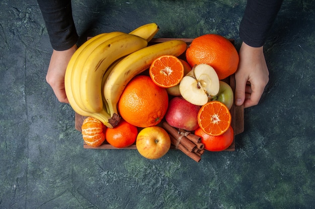 Vista superior frutas frescas naranjas mandarinas manzanas plátanos y ramitas de canela en bandeja de madera en manos femeninas sobre fondo oscuro