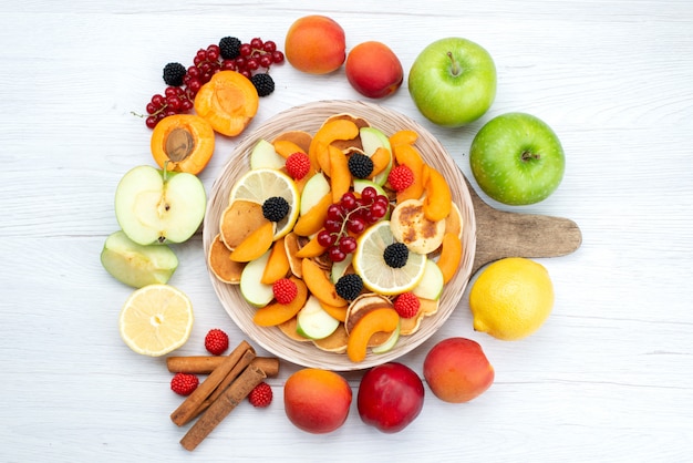 Una vista superior de frutas frescas en rodajas coloridas y maduras con frutas enteras y canela en el escritorio de madera y alimentos de color de frutas de fondo blanco