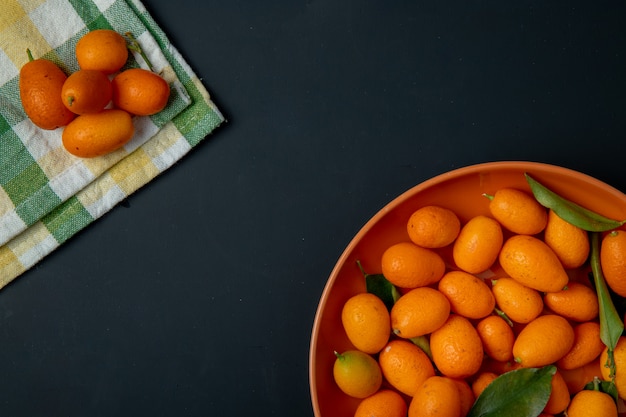 Vista superior de frutas kumquat maduras frescas en un plato en negro con espacio de copia