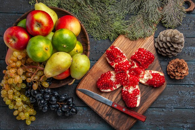 Vista superior frutas y ramas uvas blancas y negras limas peras manzanas en un tazón junto al cuchillo de granada pilled en el tablero de la cocina y ramas de abeto con conos en la mesa oscura