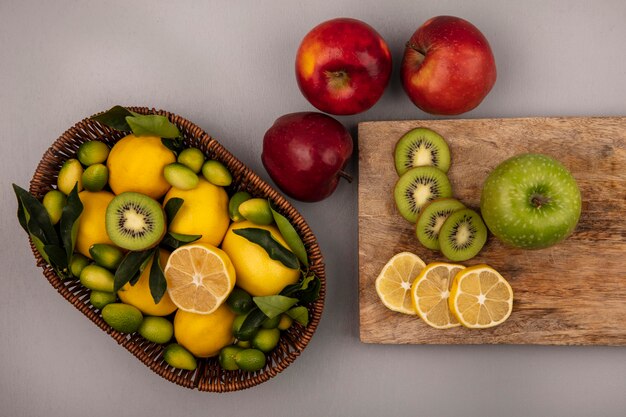 Vista superior de frutas ricas en vitaminas como kiwi kinkans y limones en un balde con rodajas de limón y kiwi en una tabla de cocina de madera con manzanas aisladas sobre un fondo gris