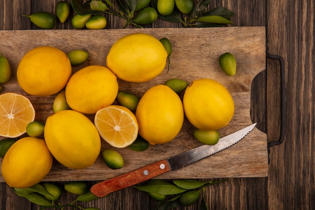 Vista superior de frutas saludables como kinkans y limones en una tabla de cocina de madera con un cuchillo sobre una superficie de madera