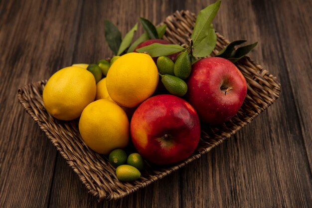 Vista superior de frutas saludables como manzanas, limones y kinkans en una bandeja de mimbre sobre una superficie de madera