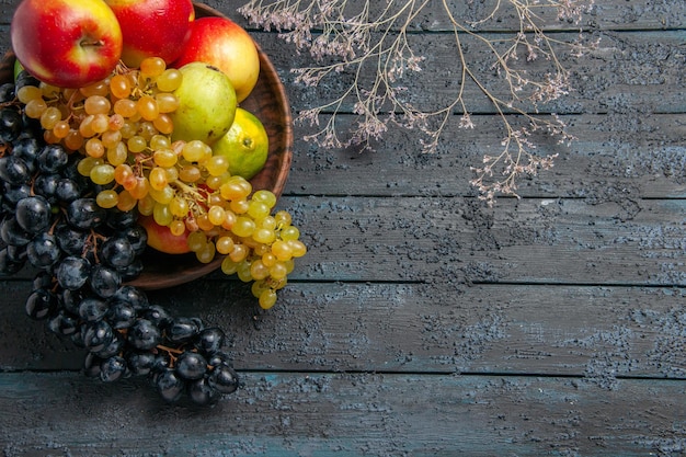 Vista superior de frutas en tazón de fuente de uvas blancas y negras limas peras manzanas junto a ramas en superficie oscura