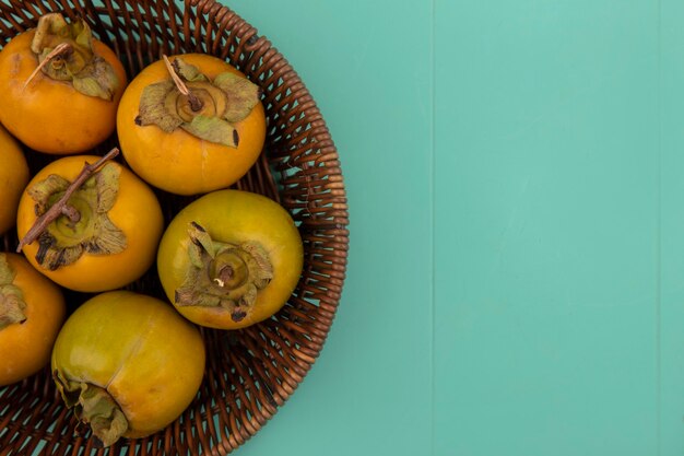 Vista superior de frutos de caqui inmaduros de color naranja en un balde sobre una mesa de madera azul con espacio de copia