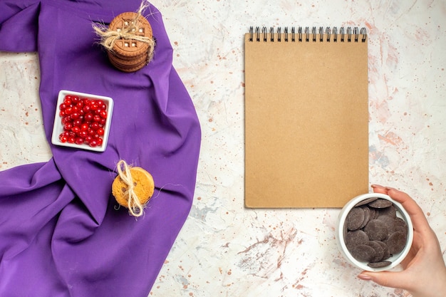 Vista superior de frutos rojos en un tazón de galletas atadas con una cuerda en el tazón de chocolate de Bloc de notas de mantón púrpura en la mano de la mujer en la mesa blanca