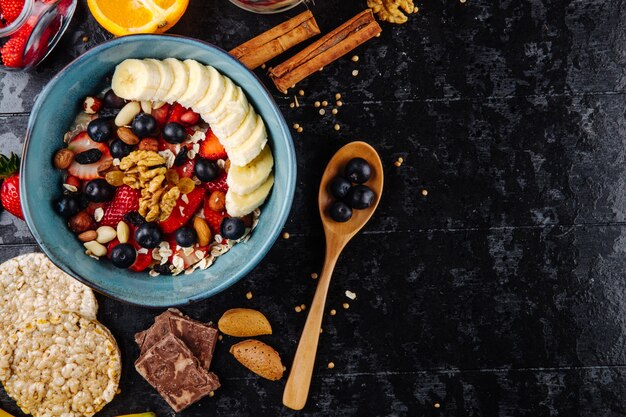 Vista superior de gachas de avena con fresas, arándanos, plátanos, frutas secas y nueces en un tazón de cerámica y cuchara de madera con bayas sobre fondo negro con espacio de copia