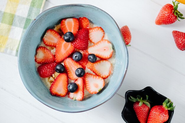 Vista superior de gachas de avena con rodajas de fresas y arándanos en un tazón de cerámica y avena durante la noche en un frasco de vidrio sobre la mesa