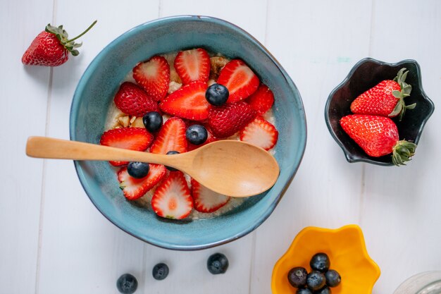 Vista superior de gachas de avena con rodajas de fresas y arándanos en un tazón de cerámica sobre superficie de madera blanca