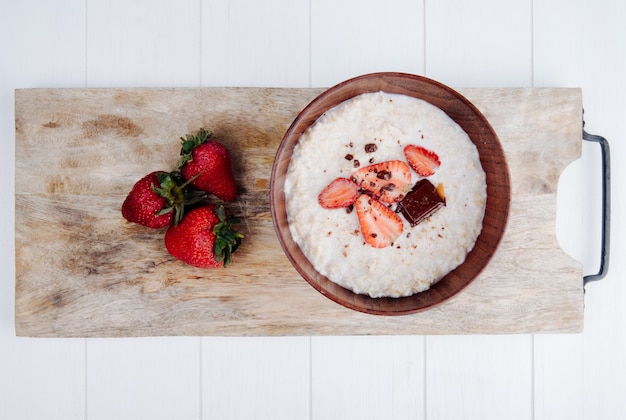 Vista superior de gachas de avena en un tazón de madera con fresas maduras frescas sobre tabla para cortar madera en rústico