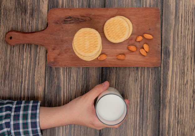 Vista superior de galletas y almendras en tabla de cortar y mano masculina con vaso de leche en la mesa de madera